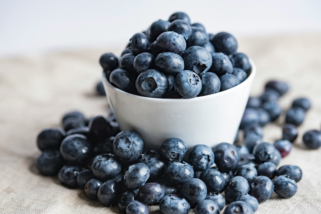 A white bowl filled with fresh blueberries, with more blueberries scattered around it on a beige fabric surface.