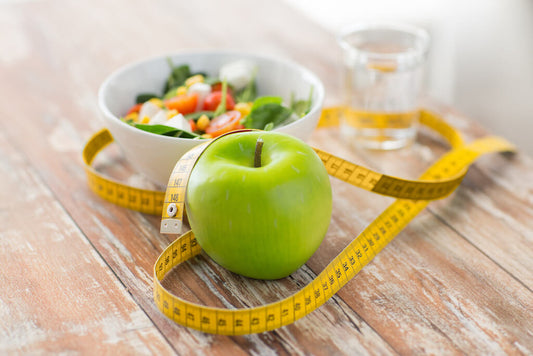 A green apple, a yellow tape measure, a bowl of salad and a glass of water are on a wooden table.