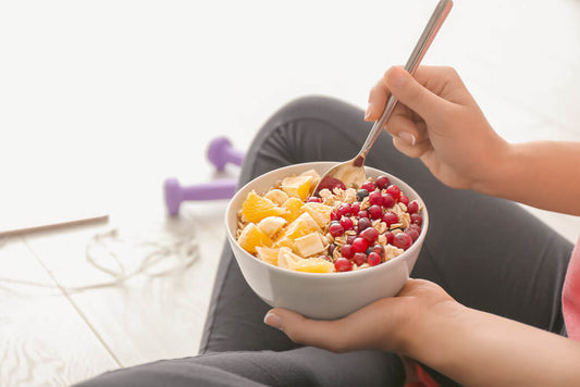 A person holds a bowl of oatmeal with orange slices, banana, and red berries - and enjoys this warming breakfast while sitting on the floor next to purple dumbbells, perfect for those who want to lose weight.
