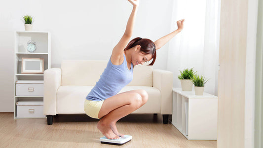 In a bright living room with a white sofa and green plants, a woman sits on a bathroom scale and stretches her arms up in joy.