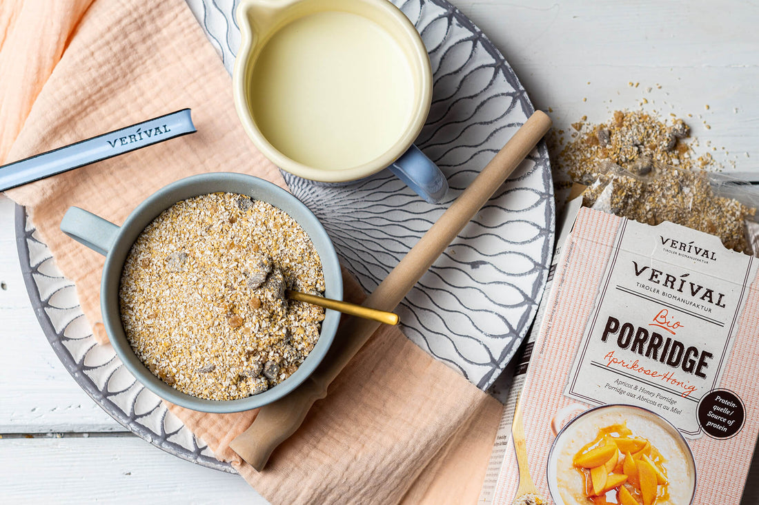 A bowl of porridge with a spoon, a jug of milk, a pack of Verival apricot and honey porridge and some spilled porridge on a tray with a napkin.