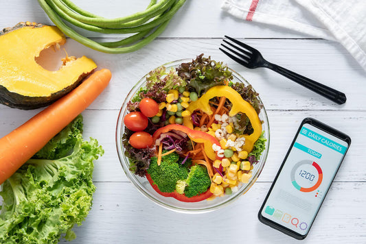 A bowl of mixed salad and vegetables next to a smartphone with a daily calorie counter, placed on a white wooden table with fresh fruits and vegetables around it.