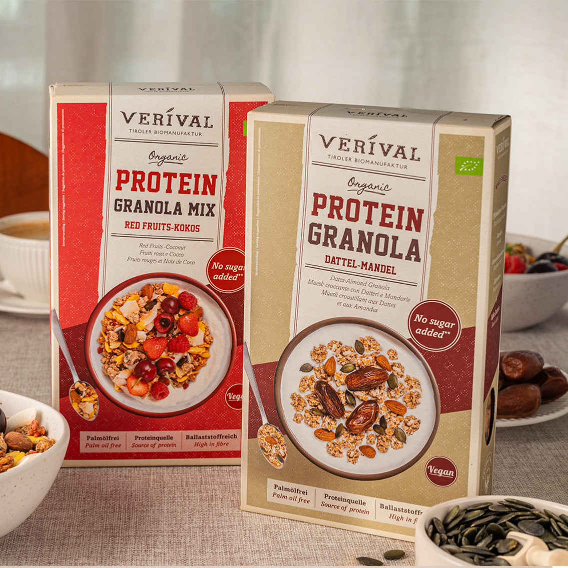 Two boxes of Verival Organic Protein Granola, one with red fruits and coconut and one with dates and almonds, are on a table with granola bowls and ingredients.