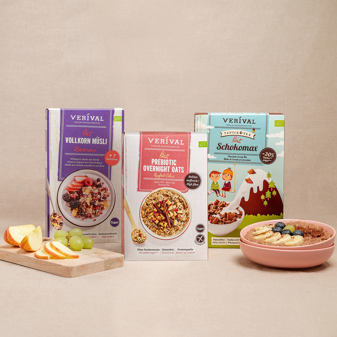 Three Verival cereal boxes are on display, including Whole Grain Muesli, Prebiotic Overnight Oats, and Schokomax. In front of them are a bowl of muesli and sliced fruit on a cutting board.