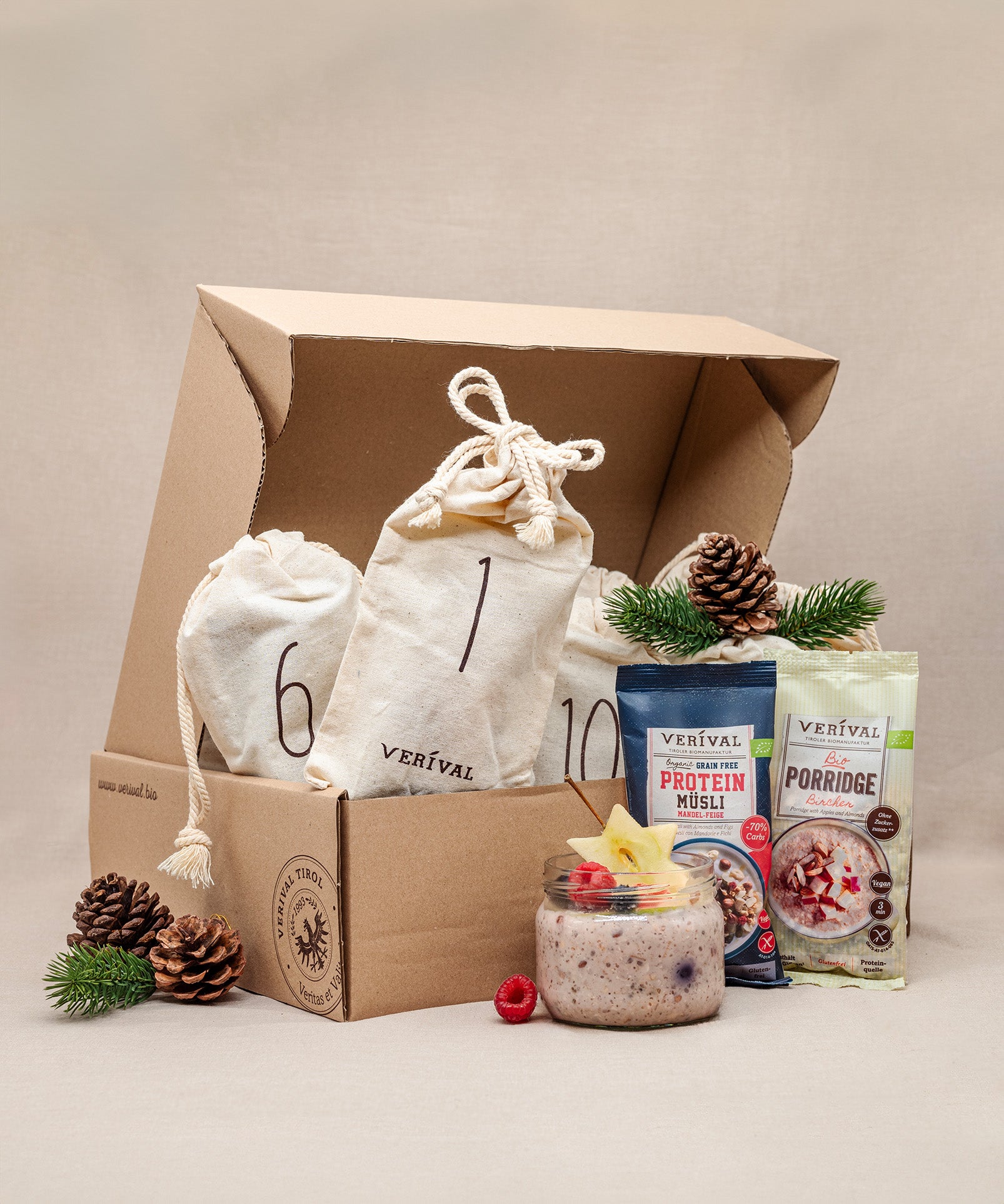 In einem geöffneten Karton, umgeben von kunstvoll arrangierten Tannenzapfen und Zweigen, befinden sich zwei Stoff-Adventskalender-Säckchen mit den Nummern 6 und 10, daneben Protein-Müsli- und Porridge-Päckchen von Verival Bio und ein Glas Himbeer-Overnight-Oats. Alle Produkte sind bio und palmölfrei.