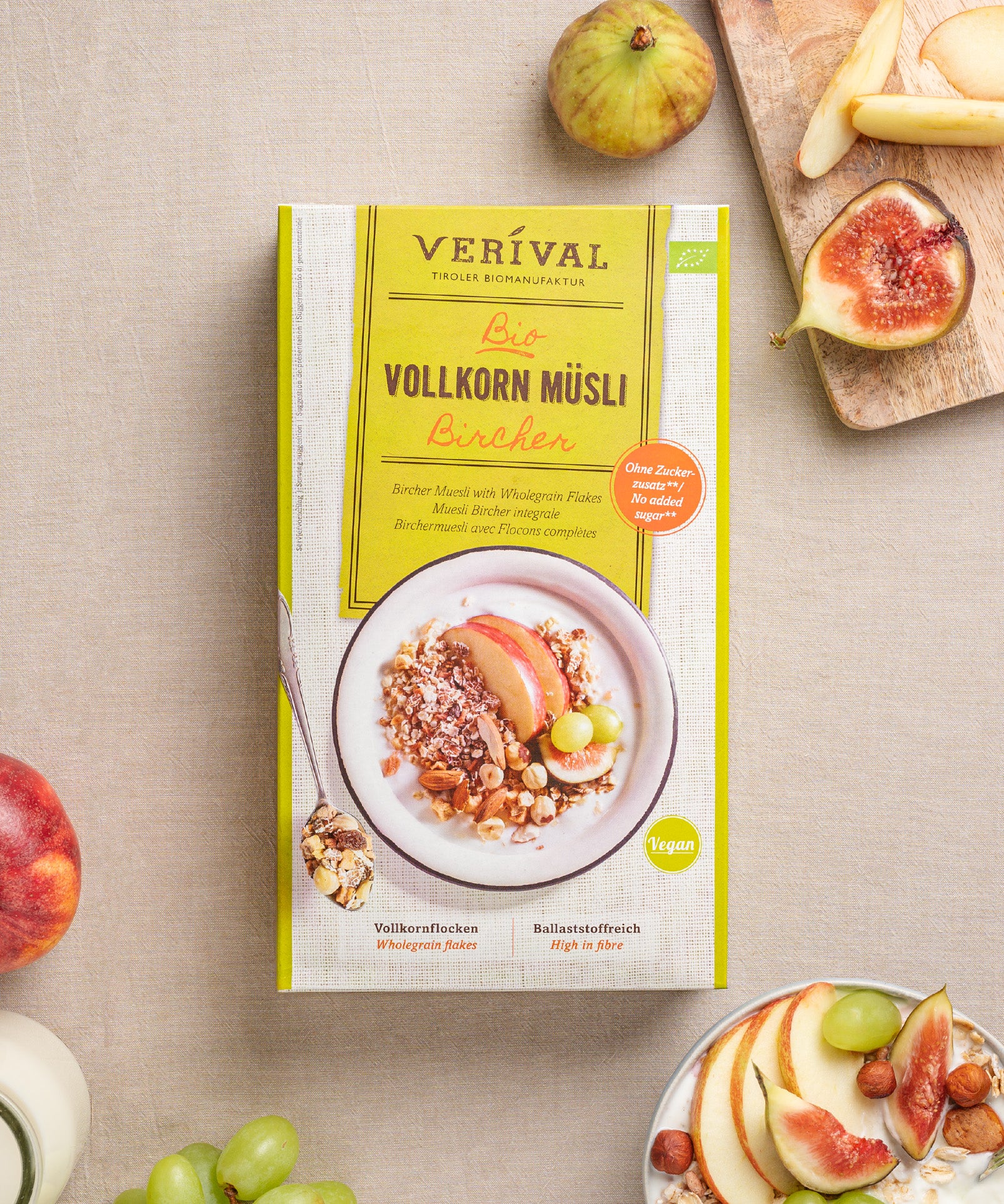 Eine Packung Bircher Vollkorn Müsli von Verival Bio steht auf einem Tisch, umgeben von Feigen, Apfelscheiben, frischen Trauben und einer Schüssel Müsli. Dieser vegane Leckerbissen steckt voller Ballaststoffe für einen guten Start in den Tag.