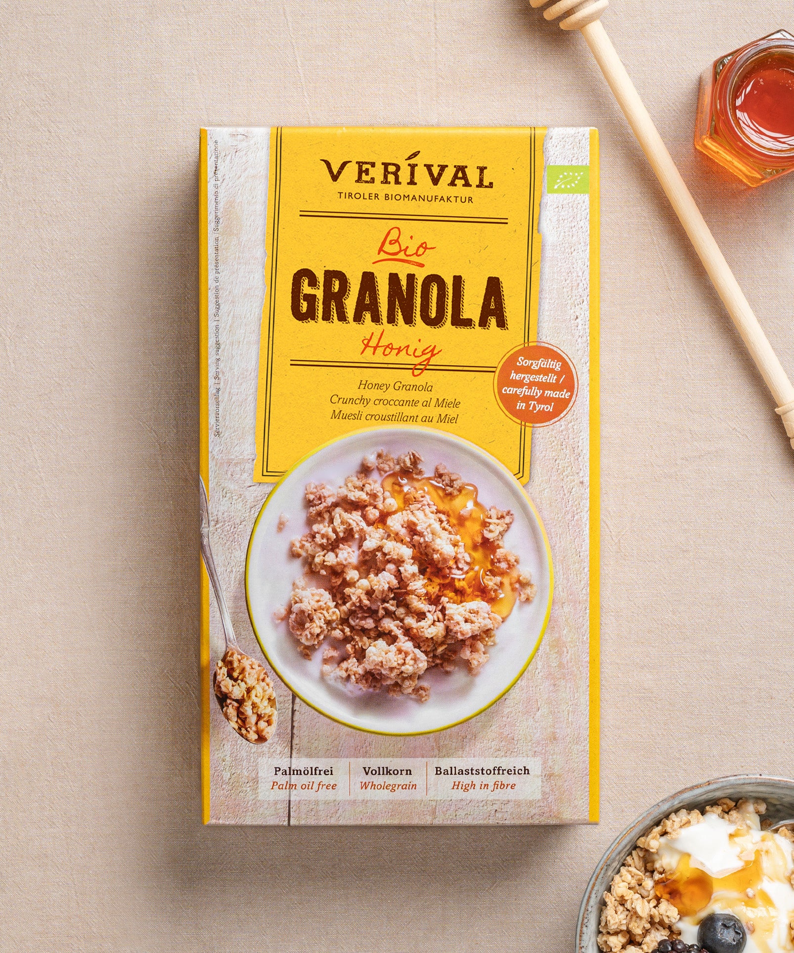 Eine Packung Honig-Granola von Verival Bio neben einer Schale knusprigem Granola mit Joghurt, dazu ein Honiglöffel und ein Glas Honig. Die Verpackung betont Bio-Zutaten, hohen Ballaststoffgehalt und wertvolles Protein für einen gesunden Start in den Tag.