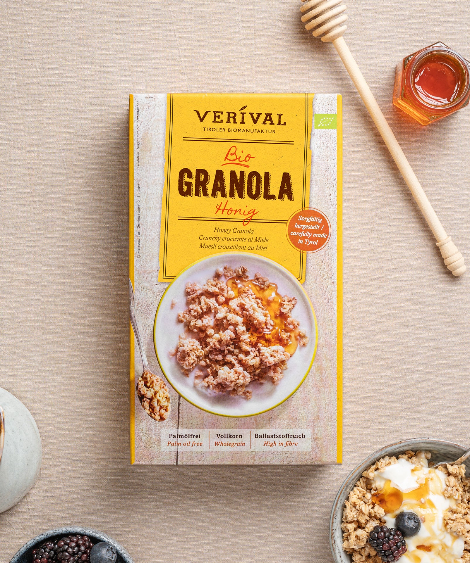 Das Honig-Granola von Verival Bio, daneben ein Löffel, ein Glas Honig und einen Honiglöffel. Dieses Produkt ist reich an Proteinen und Ballaststoffen für einen nahrhaften Start in den Tag.