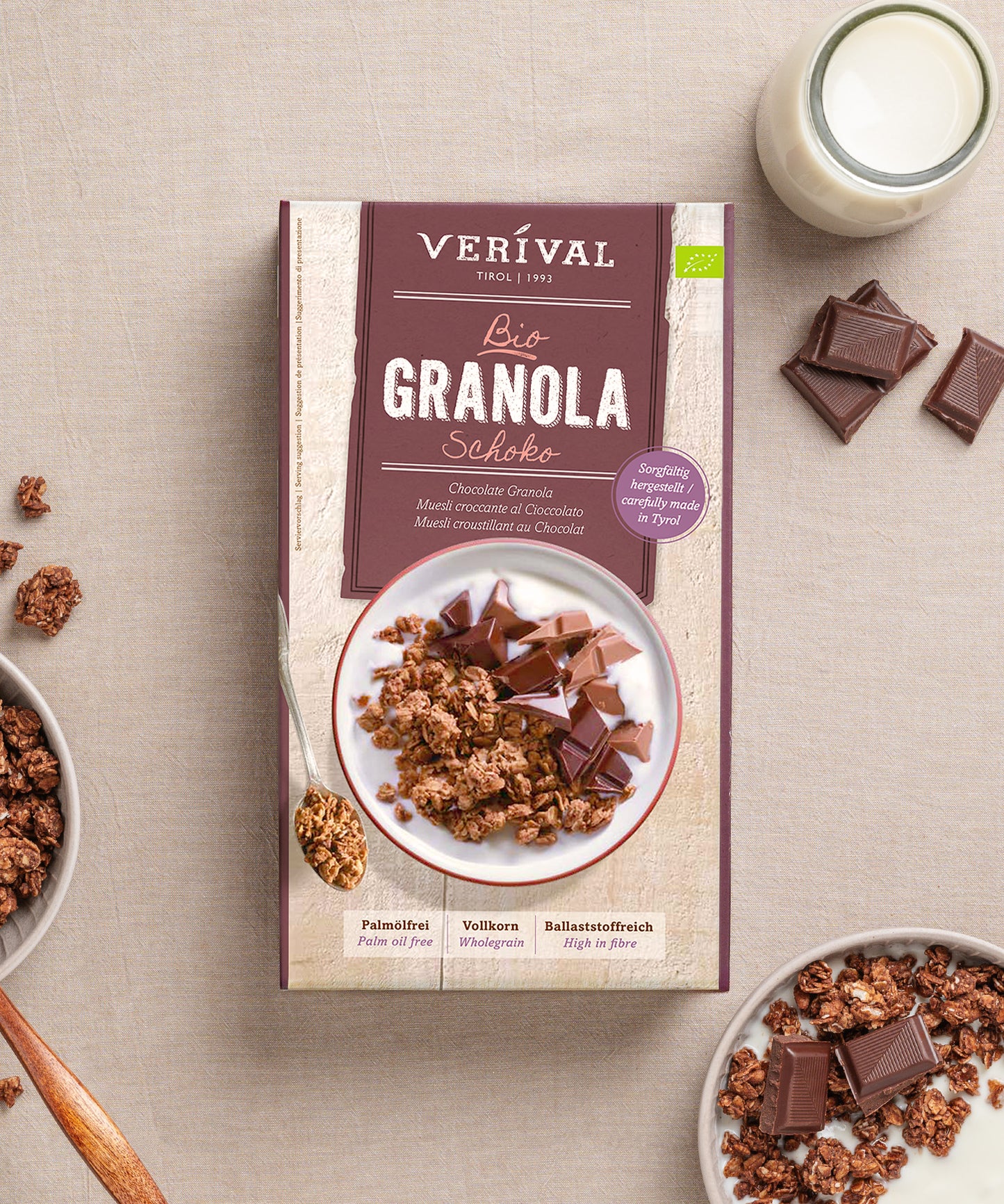 Auf einem beigen Untergrund liegt eine Schachtel Schoko Granola der Verival Bio GmbH mit knusprigen Schokostückchen, einer Schale ballaststoffreichem Granola, einem Löffel und einem Glas Milch – herrlich palmölfrei.