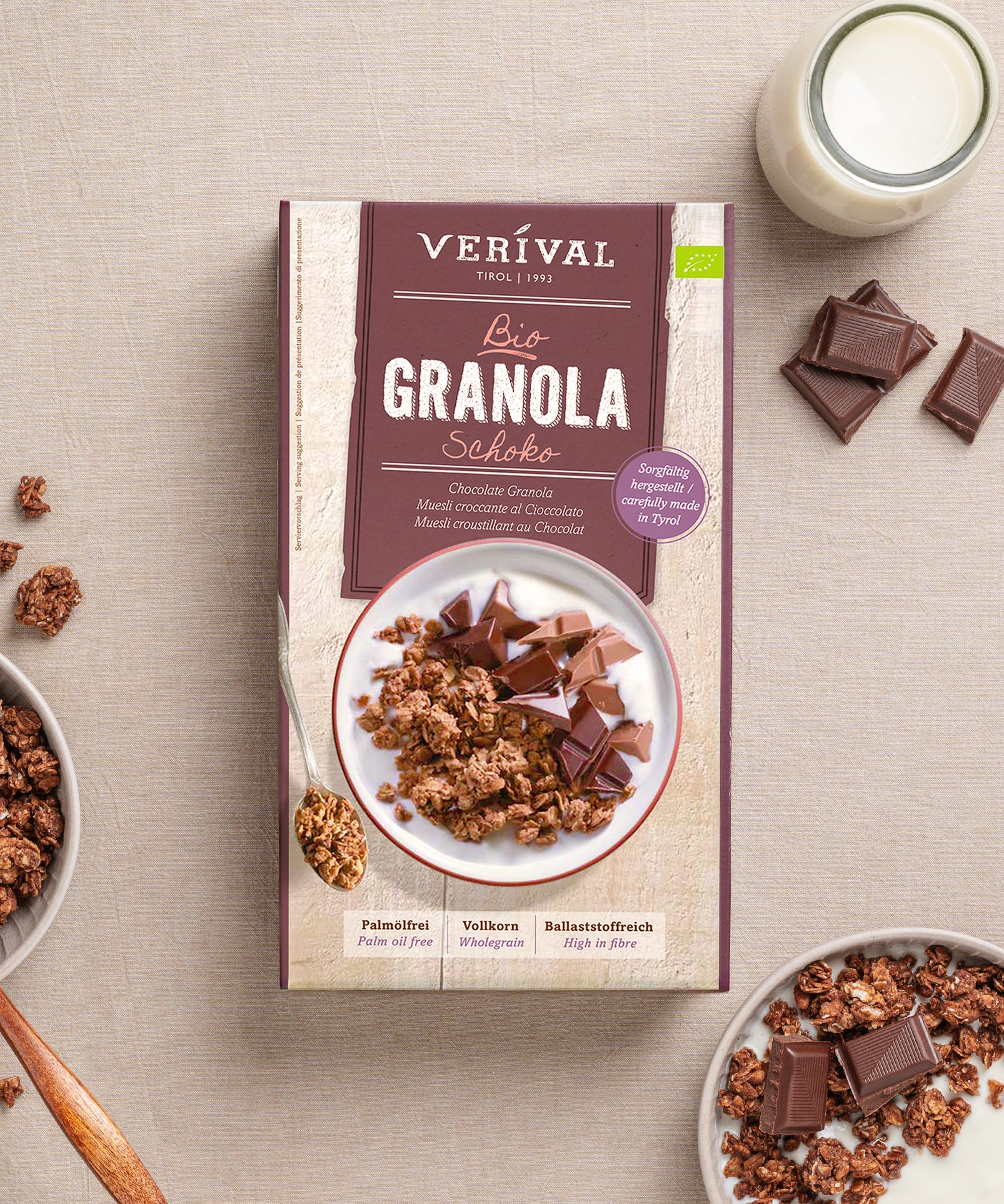 Auf einem beigen Untergrund liegt eine Schachtel Schoko Granola der Verival Bio GmbH mit knusprigen Schokostückchen, einer Schale ballaststoffreichem Granola, einem Löffel und einem Glas Milch – herrlich palmölfrei.