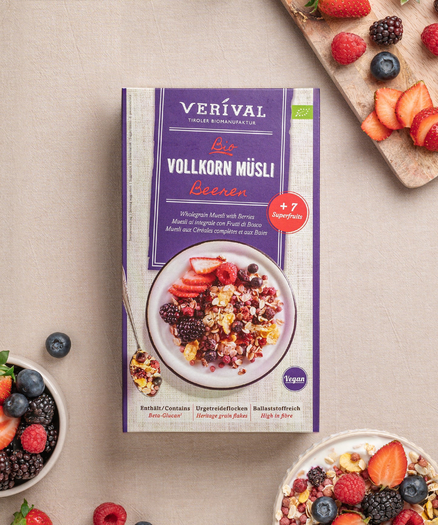 Die Beeren Vollkorn Müsli-Box knusprige Frühstücksoption steht auf einem beigen Untergrund neben einer Schüssel und einem Schneidebrett, belegt mit verschiedenen Beeren und Müsli. Die Verpackung erwähnt den veganen Charakter, den ballaststoffreichen Gehalt und die Verwendung von +7 Superfrüchten.