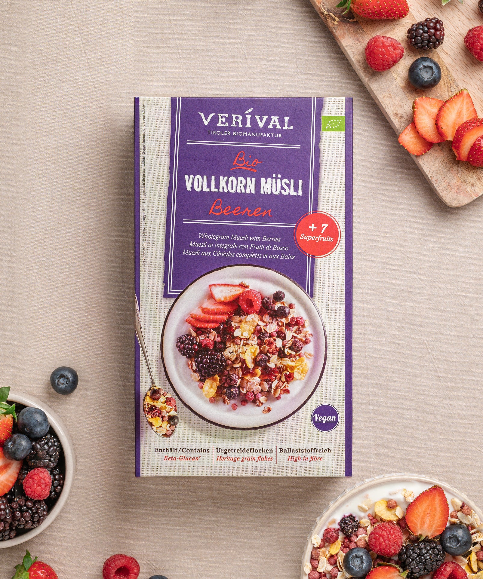 Die Beeren Vollkorn Müsli-Box knusprige Frühstücksoption steht auf einem beigen Untergrund neben einer Schüssel und einem Schneidebrett, belegt mit verschiedenen Beeren und Müsli. Die Verpackung erwähnt den veganen Charakter, den ballaststoffreichen Gehalt und die Verwendung von +7 Superfrüchten.