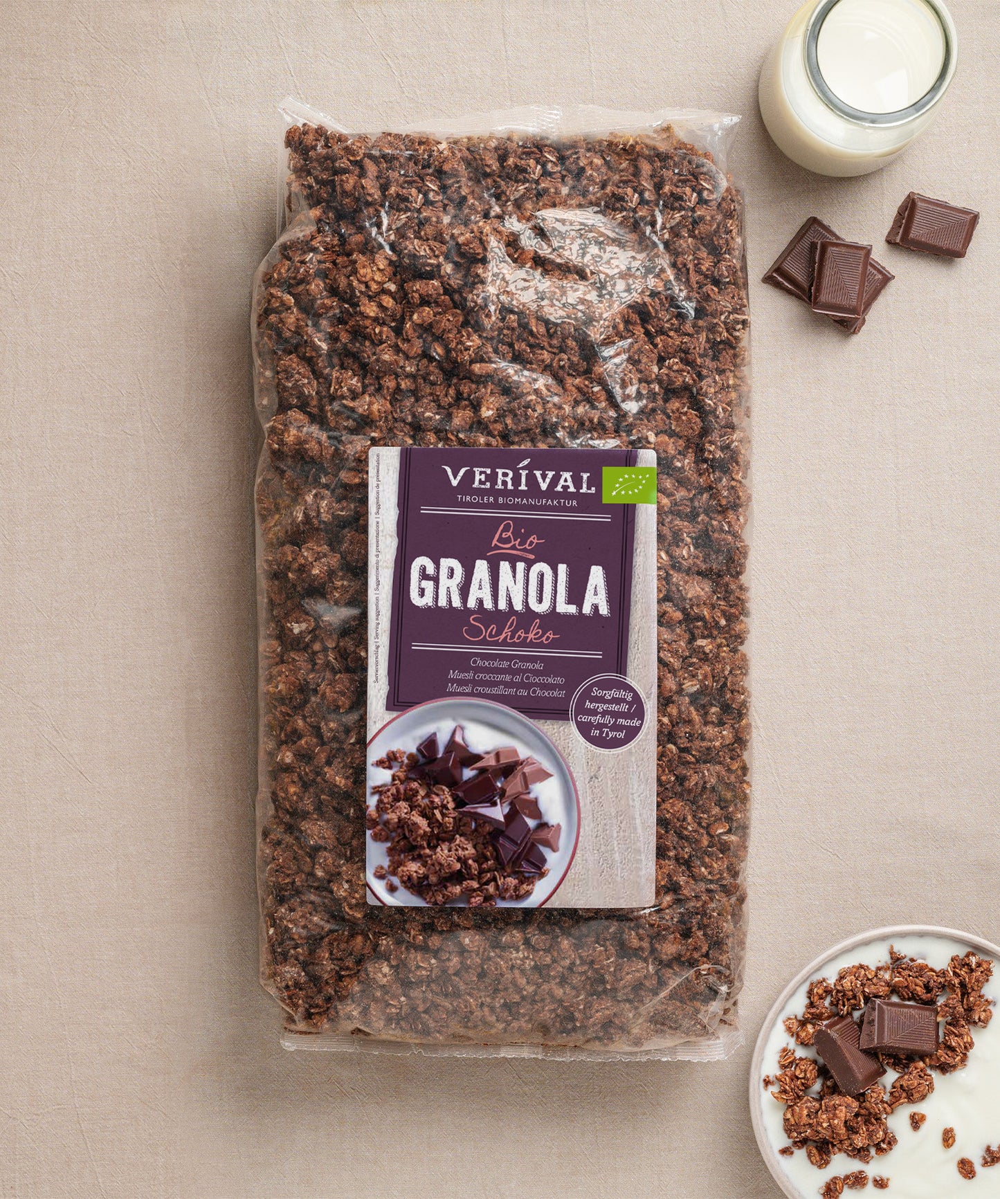 Auf einem beigen Untergrund liegt eine Packung Schoko Granola von Verival Bio, umgeben von Schokoladenstückchen, einem kleinen Milchkännchen und einem Teller mit einer knusprigen Portion des palmölfreien Granolas.