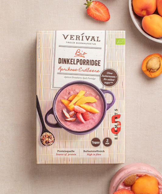 Eine Schachtel Aprikosen-Erdbeer Dinkelporridge von Verival Bio steht neben einer Schüssel mit demselben Porridge, garniert mit frischem Obst und umgeben von Aprikosen – ein köstliches TCM-Frühstück mit hohem Ballaststoffgehalt.