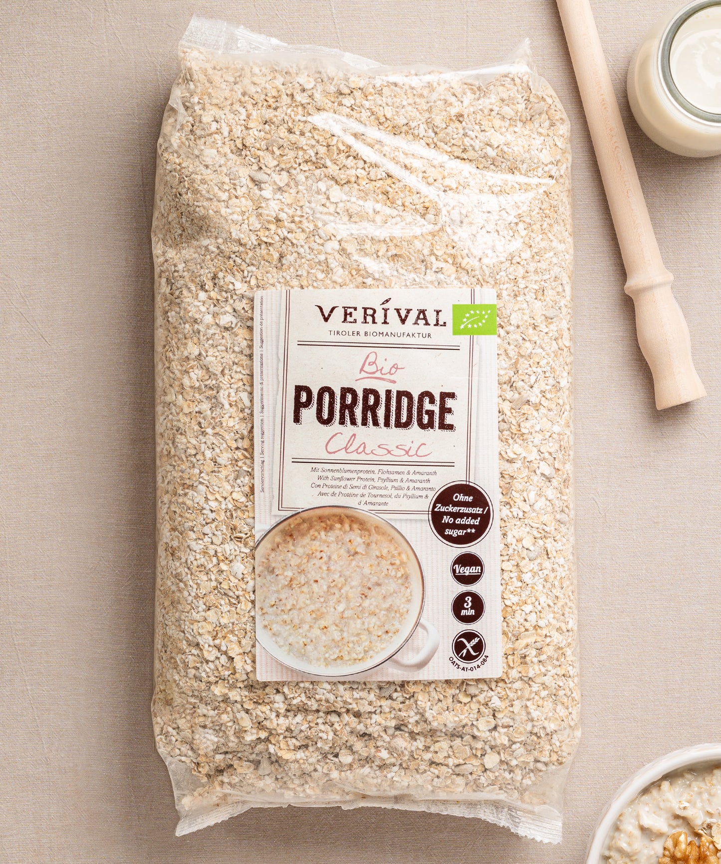 Das Classic Porridge von Verival Bio präsentiert sich mit einer Schüssel Porridge, einem Holzlöffel und einem Glas Milch auf beigefarbenem Grund. Diese glutenfreie Option ist ballaststoffreich und bietet einen nahrhaften Start in den Tag.