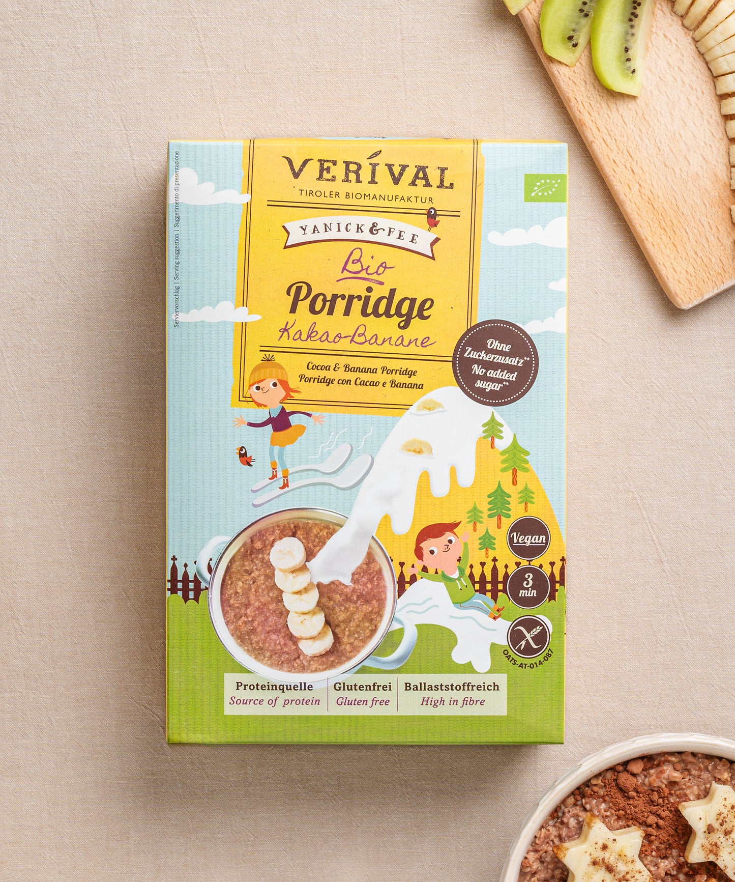 Eine Packung ballaststoffreicher Kids Porridge Kakao-Banane von Verival Bio steht auf einem hellen Untergrund neben einer Schüssel glutenfreiem Porridge mit Bananenscheiben und Kiwischeiben.