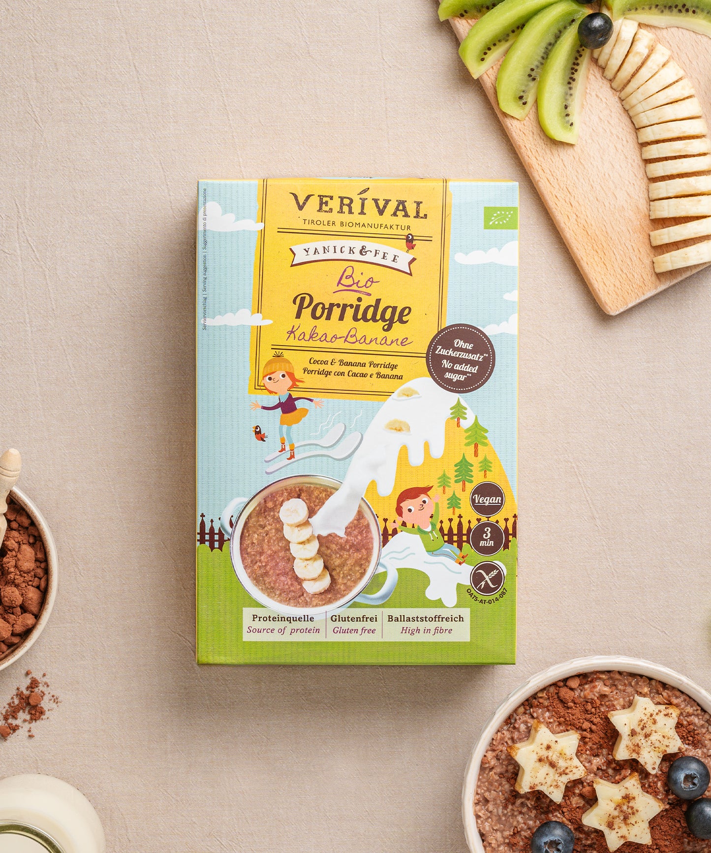 Eine Packung Verival Bio Kinderporridge Kakao-Banane steht neben einer Schüssel cremigem Porridge mit Kiwi- und Bananenscheiben sowie einer Kokosnussschale mit Nüssen auf dem Tisch. Dieser glutenfreie Genuss steckt voller Ballaststoffe für einen nahrhaften Start in den Tag.