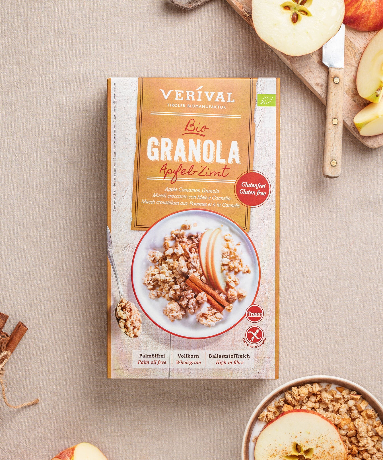Eine Packung Apfel-Zimt-Granola von Verival Bio mit einer Schüssel proteinreichem, glutenfreiem und veganem Knusper-Frühstück-Granola und geschnittenen Äpfeln auf dem Tisch. Frische Äpfel auf einem Schneidebrett sorgen für zusätzliche Ballaststoffe.