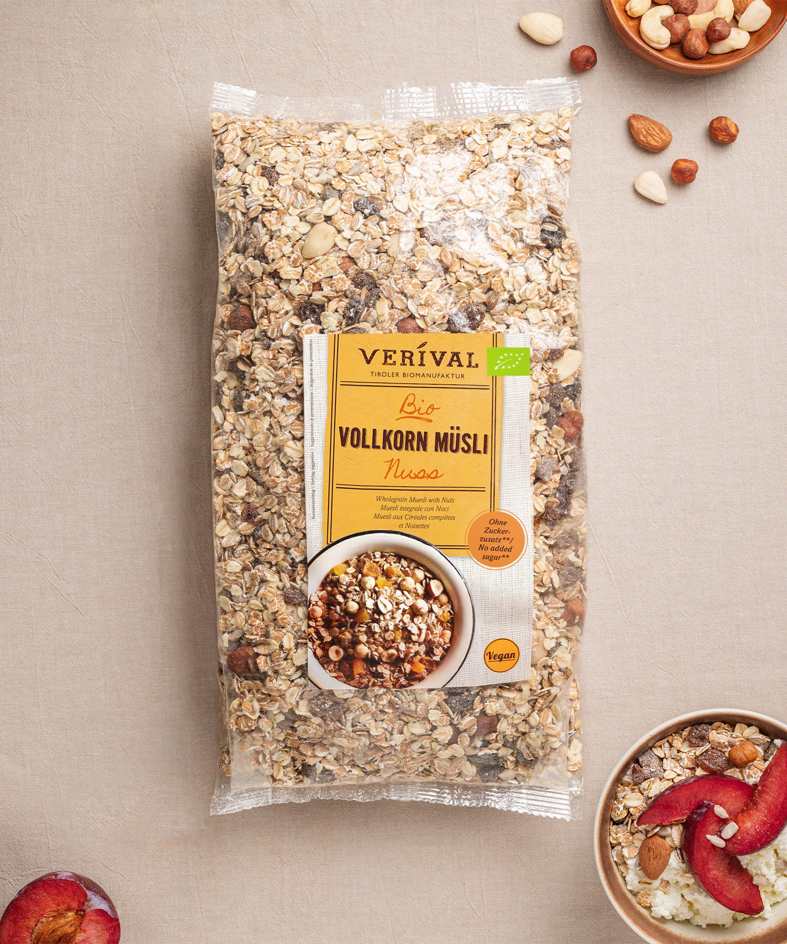 Auf einer beigen Fläche steht eine Packung Nuss-Vollkorn-Müsli von Verival Bio mit verstreuten Nüssen und einer kleinen Müslischale, garniert mit roten Fruchtscheiben.