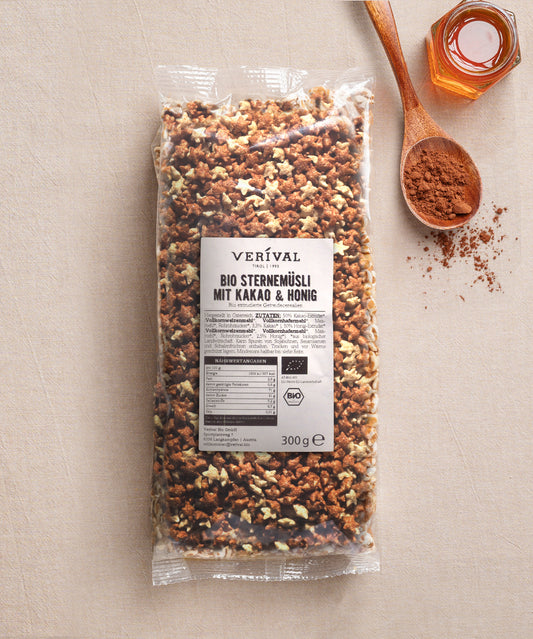 Eine 300g-Packung Verival Bio GmbH Bio Sternemüsli mit Kakao & Honig steht auf einer beigen Fläche, daneben ein Holzlöffel mit Kakaopulver und ein kleines Glas Honig.