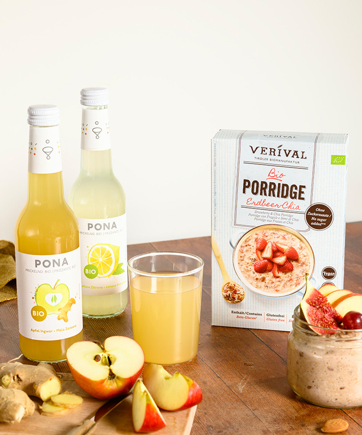Zwei Flaschen PONA Primofiore Zitrone und eine Packung Erdbeer-Chia Porridge ergänzt mit geschnittenen Äpfeln und Ingwer. Daneben steht ein kleines Glas mit Porridge auf einer hellen Holzoberfläche.
