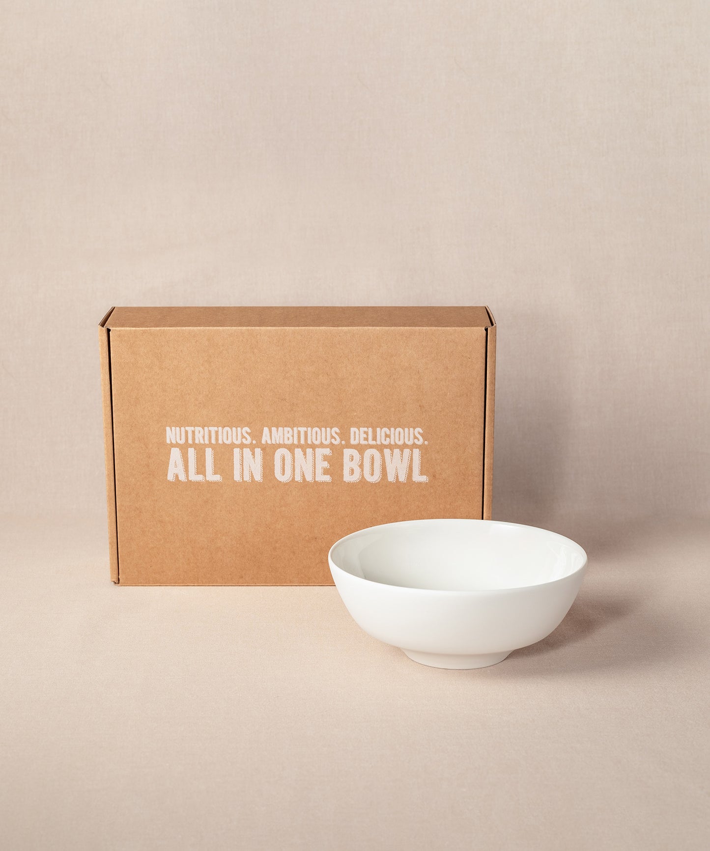 Eine braune Kartonschachtel mit dem Text „Nutritious. Ambitious. Delicious. All in one bowl“ steht aufrecht,  neben einer eleganten, weißen Porridge-Schüssel aus Keramik. Die Schüssel ist Bestandteil des Sets „Breakfast Lover“ von Verival Bio.