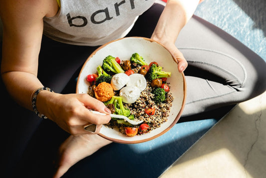 A person in workout clothes sits cross-legged, holding a bowl of quinoa, broccoli, cherry tomatoes, and poached eggs, and eating with a fork.