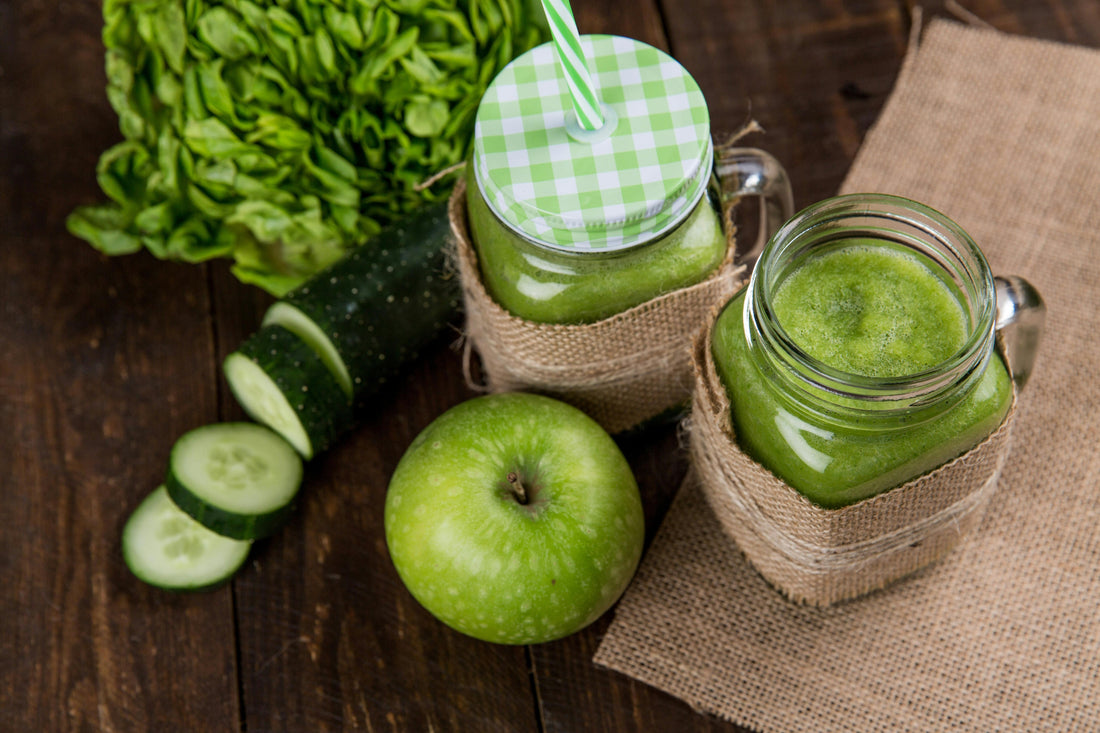 Two mason jars of green smoothie on burlap, with green apple, cucumber slices, and lettuce on a wooden table.