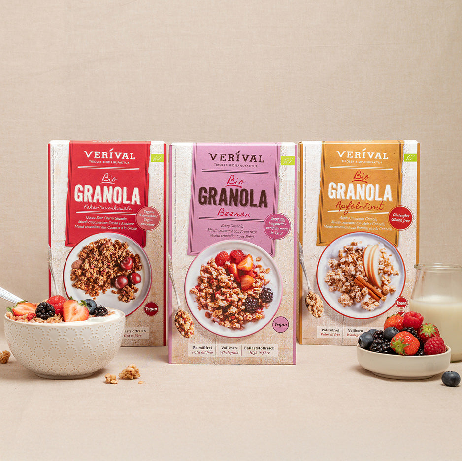Drei Schachteln Verival-Granola (Classic, Beeren, Apfel-Zimt) stehen aufrecht, im Vordergrund eine Schale mit Granola und frischen Beeren sowie ein Glas Milch.