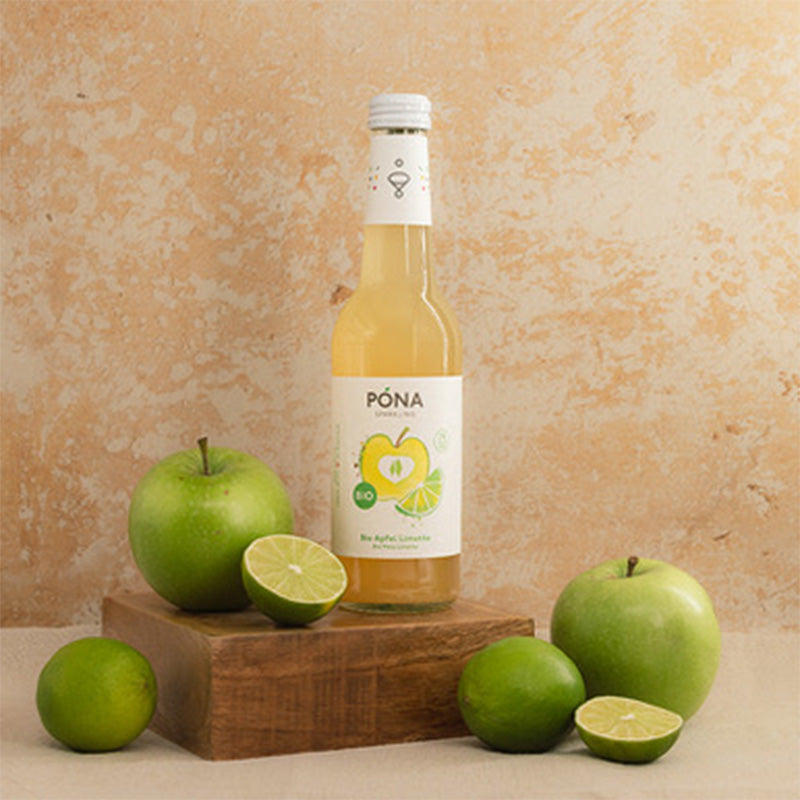 Eine Flasche PONA Apfel-Limetten-Getränk auf einem Holzblock, umgeben von grünen Äpfeln und Limettenscheiben, vor einem strukturierten beigen Hintergrund.