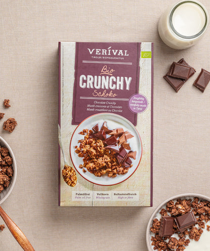 Eine Schachtel aus dem Set "Best Breakfast Friends" der Verival Bio GmbH steht auf einer beigen Fläche, umgeben von Müslischalen, Schokoladenstückchen und einem Glas Milch - das ideale Bio Frühstück oder Geschenkset für Schokoladenliebhaber.