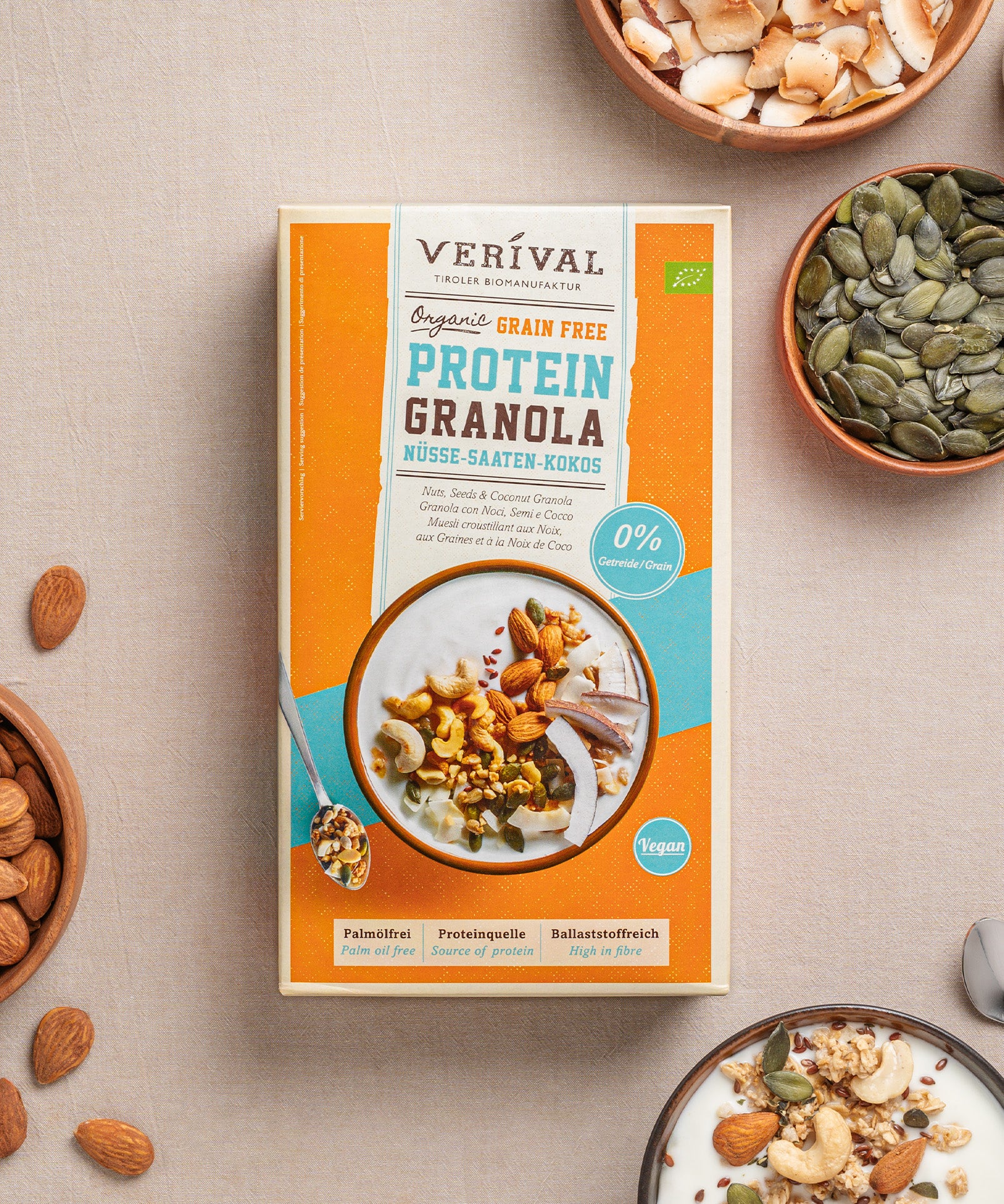 Eine Paket Grain Free Protein Granola Nüsse-Saaten-Kokos von Verival Bio, umgeben von Schalen mit Samen, Nüssen und einer mit Granola gefüllten Joghurtschale auf einer hellen Oberfläche, bietet köstliche pflanzliche Proteine für deinen Low-Carb-Lebensstil.