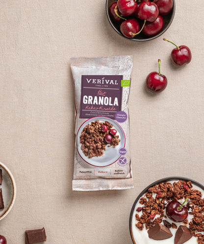 Ein verpackter Verival Bio GmbH Bio Kakao-Kirsche Granola glutenfrei Riegel liegt auf einer beigen Fläche, umgeben von Schalen mit Kirschen, einer Schale mit glutenfreiem Granola mit Kirschen und Schokoladenstücken.