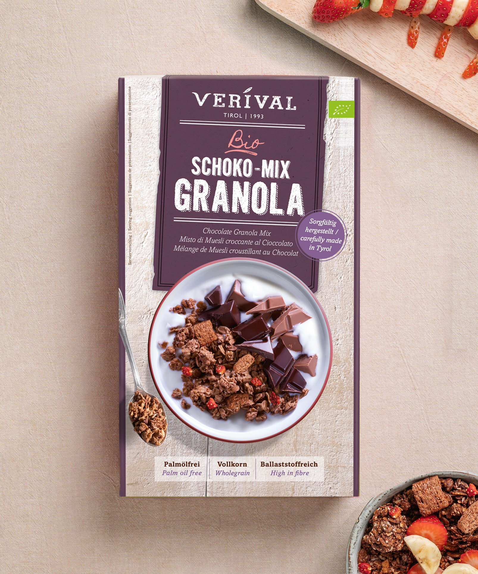 Eine Schachtel Verival Bio GmbH Bio Schoko Mix Granola steht auf einer hellen Fläche, neben einer Schüssel mit knusprigen Frühstücksflocken und einem Löffel. Die Verpackung wirbt für das ballaststoffreiche Vollkorn-Granola mit Schokostückchen und zeigt es mit Milch serviert.