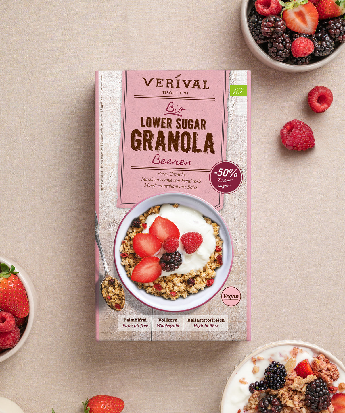 Auf einem beigen Untergrund steht eine Schachtel „Bio Lower Sugar Beeren-Müsli“ von Verival Bio GmbH, umgeben von Schalen mit Vollkornmüsli, Joghurt, gemischten Beeren sowie losen Erdbeeren, Himbeeren und Brombeeren – perfekt für einen zuckerarmen Genuss.