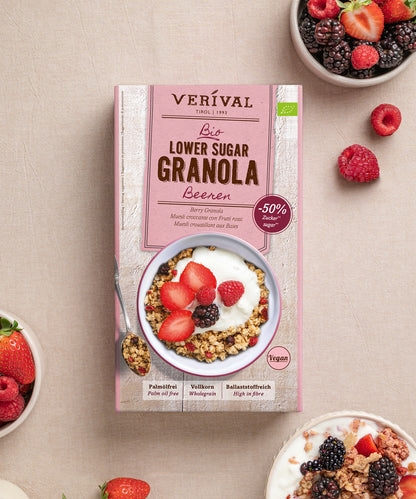 Auf einem beigen Untergrund steht eine Schachtel „Bio Lower Sugar Beeren-Müsli“ von Verival Bio GmbH, umgeben von Schalen mit Vollkornmüsli, Joghurt, gemischten Beeren sowie losen Erdbeeren, Himbeeren und Brombeeren – perfekt für einen zuckerarmen Genuss.