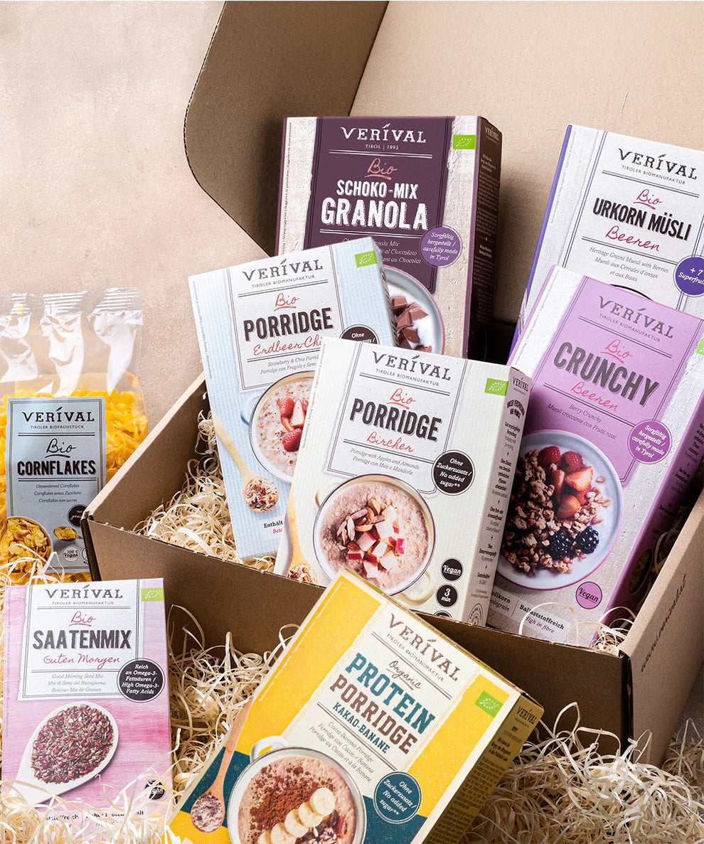 Das Set "Best Breakfast Friends" der Verival Bio GmbH zeigt eine Auswahl an Frühstücksflocken - Granola, Porridge, Müsli, Cornflakes und Körnermischung - ansprechend auf Stroh neben einer offenen Pappschachtel arrangiert.